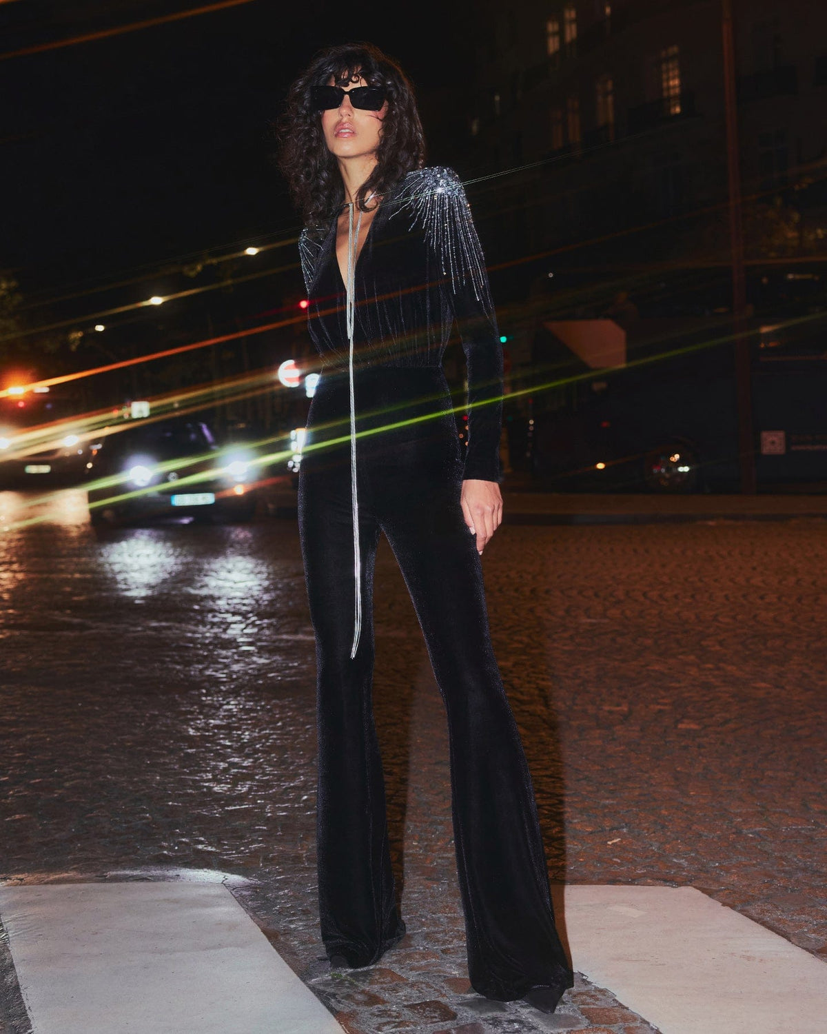 Person in a black sequined jacket and bell-bottom pants standing on a street at night.