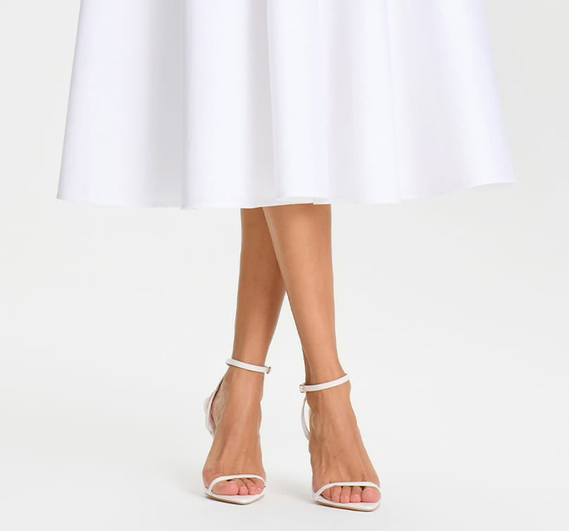 Clear high-heeled sandals worn with a white dress on a white background