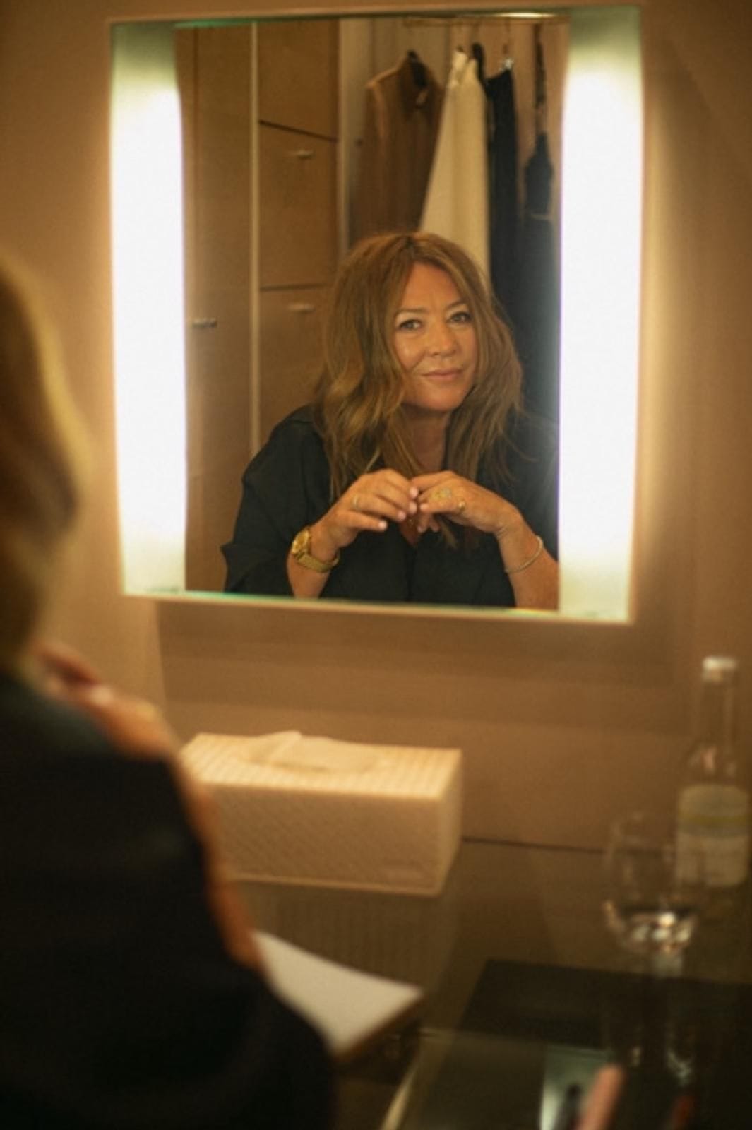 Woman looking at herself in a mirror with a lighted frame, wearing a black top and gold bracelet.